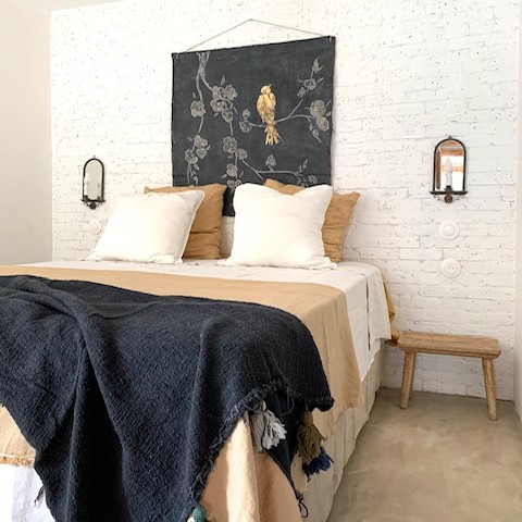Une chambre à coucher de style rustique avec un grand lit couvert d'un plaid bleu foncé, de draps beiges et blancs, et d'oreillers moelleux. Un tableau d'oiseau décore le mur en briques blanches, flanqué de deux appliques murales. Un tabouret en bois est placé à côté du lit.