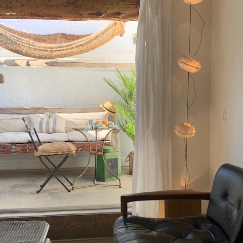 Un patio ensoleillé avec des poutres en bois apparentes, un hamac, un banc rembourré, une petite table ronde en fer forgé et une chaise pliante, visible depuis l'intérieur à travers une grande ouverture encadrée de rideaux blancs.