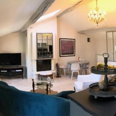 Un salon spacieux avec un canapé bleu, un tapis beige, et une cheminée moderne. Une table à manger avec des chaises blanches est visible, ainsi qu'une télévision et un lustre doré.