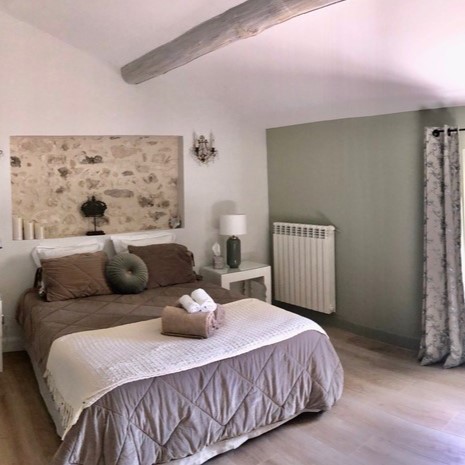Une chambre lumineuse avec un lit fait de draps marron, une tête de lit en pierre exposée avec une niche décorative, et une poutre en bois au plafond.