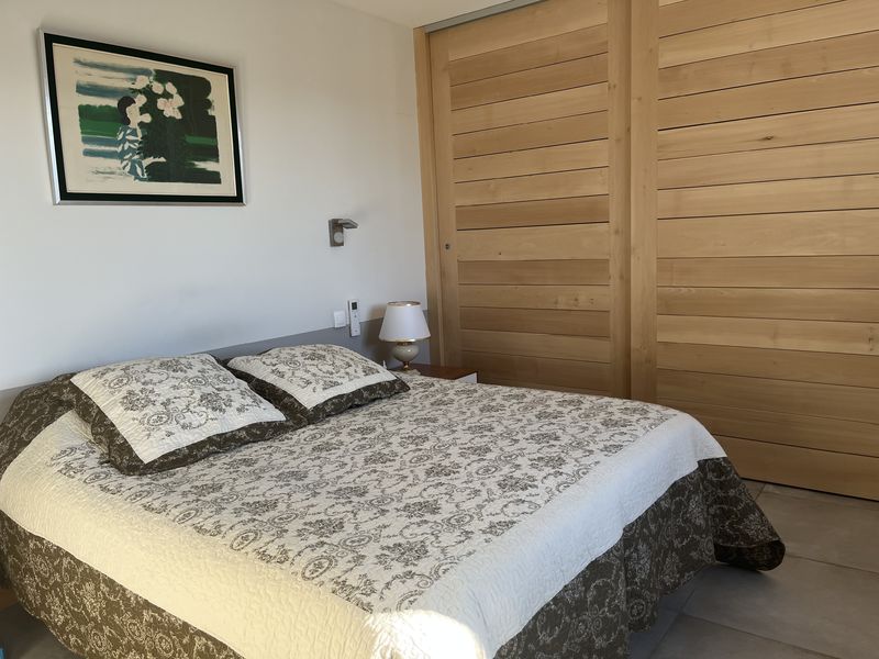 Une chambre à coucher aux murs clairs, avec un lit double couvert d'une courtepointe blanche et grise à motifs, flanqué d'une table de chevet avec une lampe. Des placards en bois clair occupent un mur entier, et un tableau abstrait est accroché au-dessus du lit.