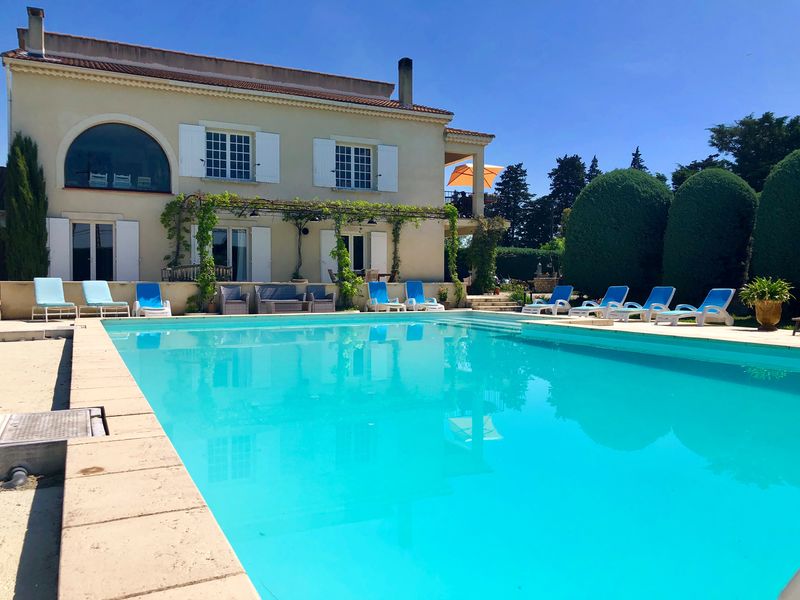 Une grande villa provençale avec des volets blancs, une piscine extérieure aux eaux bleues claires, et de nombreuses chaises longues bleues et blanches disposées autour.