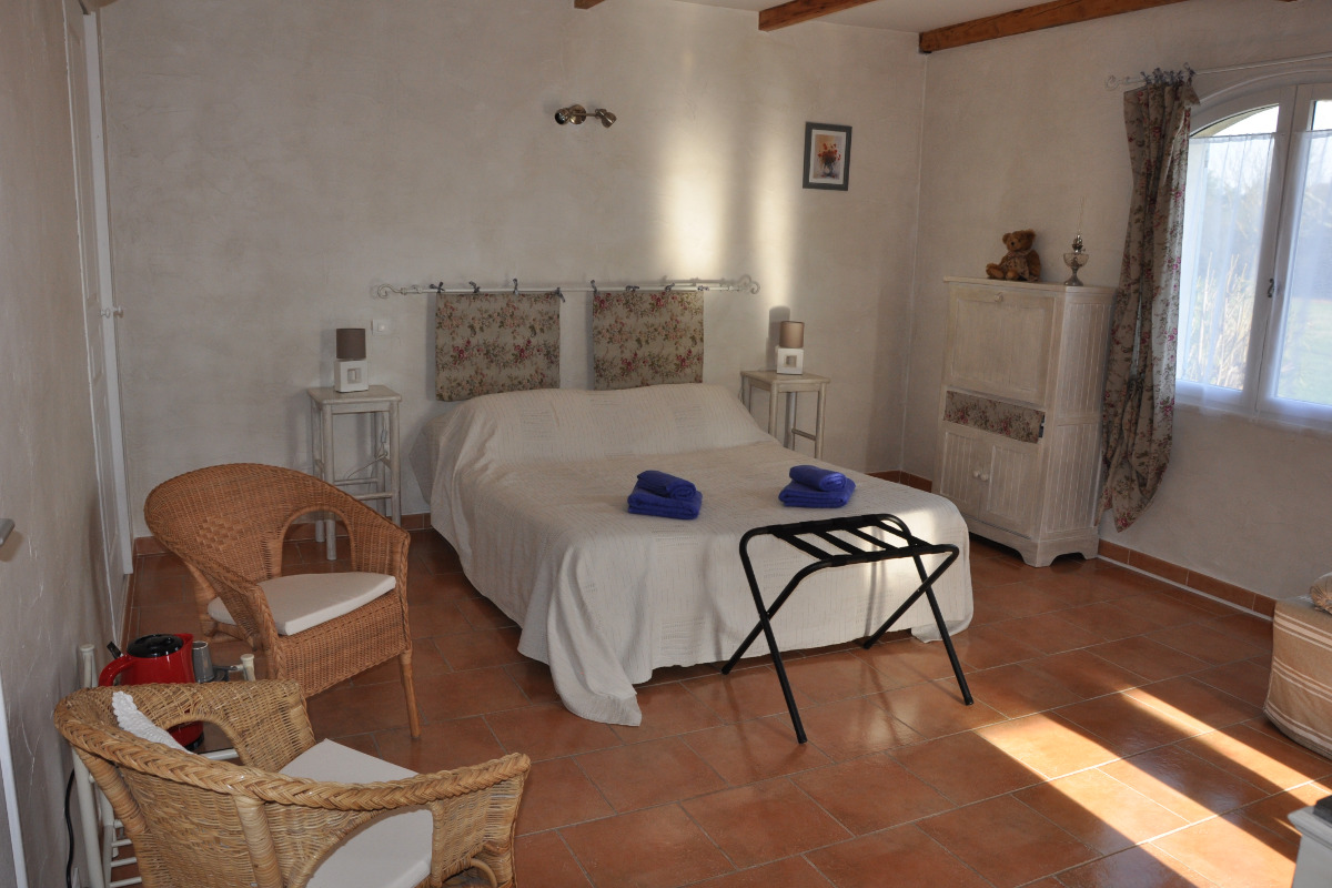 Une chambre chaleureuse avec un lit double couvert d'une couette blanche et de serviettes bleues, flanqué de tables de chevet et de lampes. Une tête de lit rembourrée à motifs floraux, un meuble blanc avec un ours en peluche, et deux fauteuils en rotin complètent la pièce.