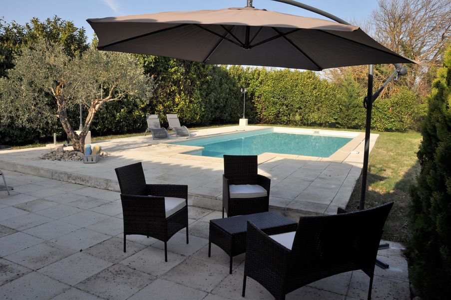 Une terrasse carrelée avec un salon de jardin en rotin noir sous un grand parasol beige. Une piscine rectangulaire est visible à l'arrière-plan, entourée de verdure et de chaises longues, avec un olivier sur la gauche.