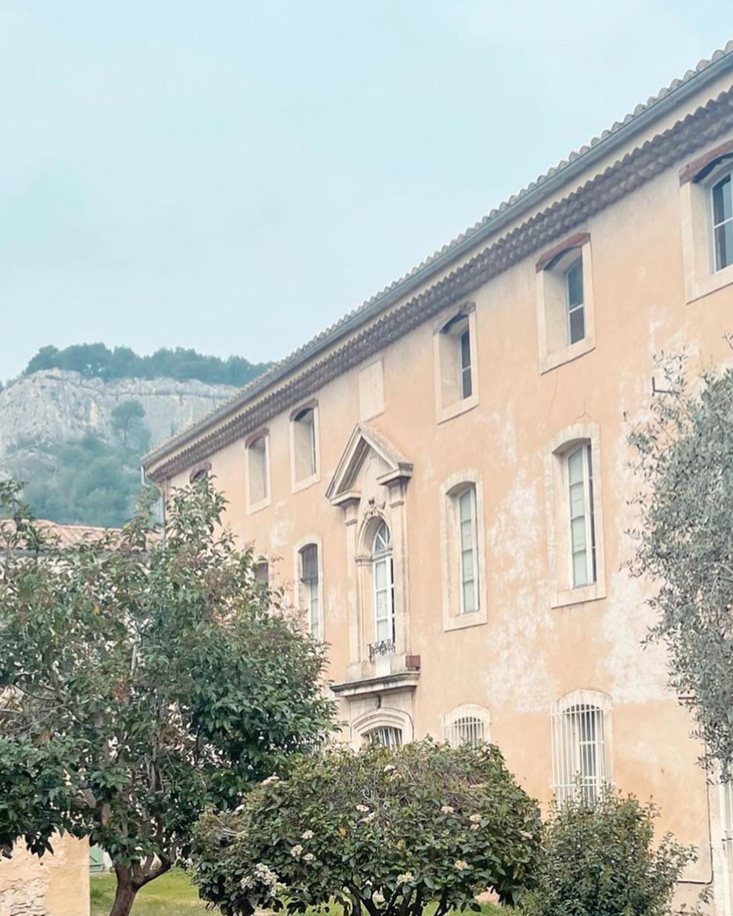 Un grand bâtiment de couleur pêche avec des fenêtres symétriques et un balcon orné au centre, bordé d'arbres feuillus et d'oliviers, sous un ciel pâle. Une colline rocheuse se dresse en arrière-plan.