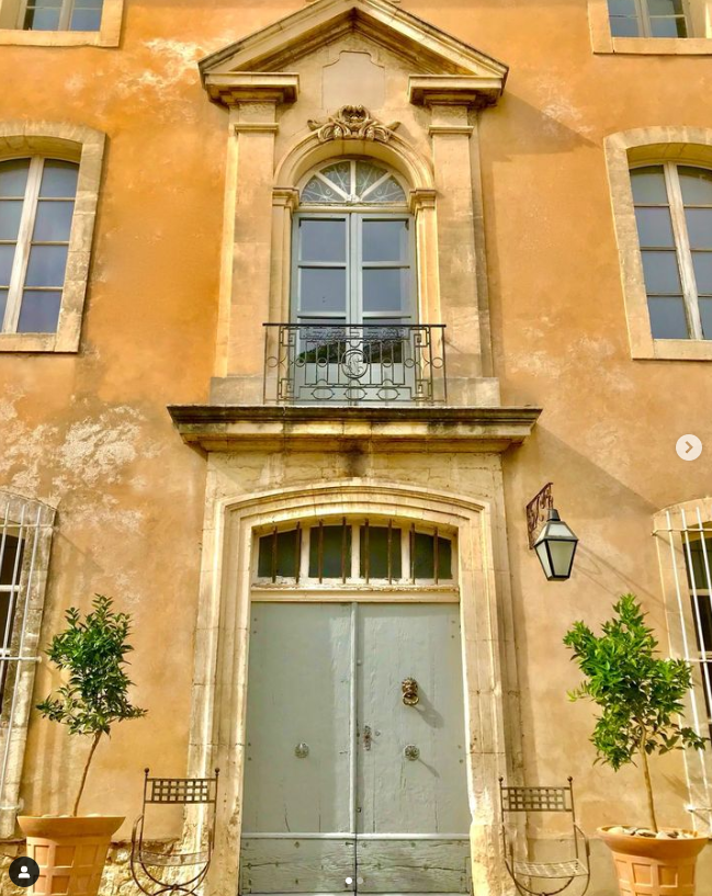 Une façade de bâtiment ocre avec une grande porte d'entrée en bois clair et un balcon orné au-dessus. Des pots de plantes et deux chaises en fer forgé sont disposés de chaque côté de la porte.