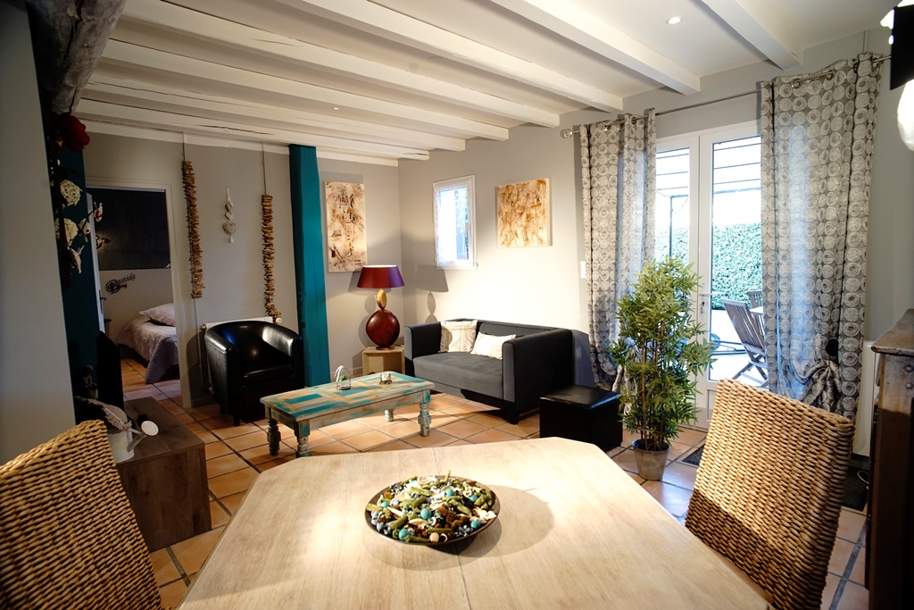 Une pièce de vie ouverte avec des poutres blanches au plafond, un sol carrelé, un salon avec canapé et fauteuils sombres, et une table basse turquoise. Une table à manger en bois avec des chaises en rotin est au premier plan, et une fenêtre donne sur une terrasse.