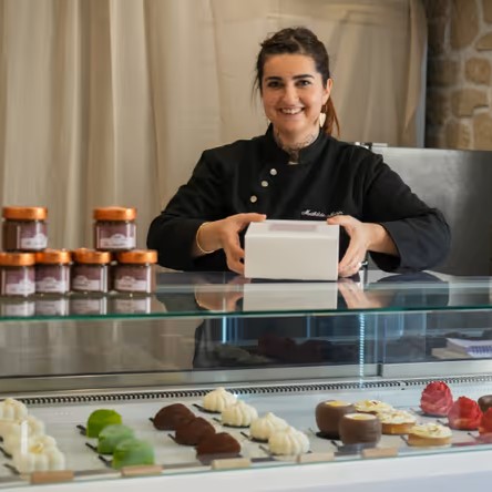 Une cheffe souriante, vêtue d'une veste de cuisine noire, tenant une boîte blanche derrière un comptoir vitré rempli de pâtisseries colorées et de petits pots.