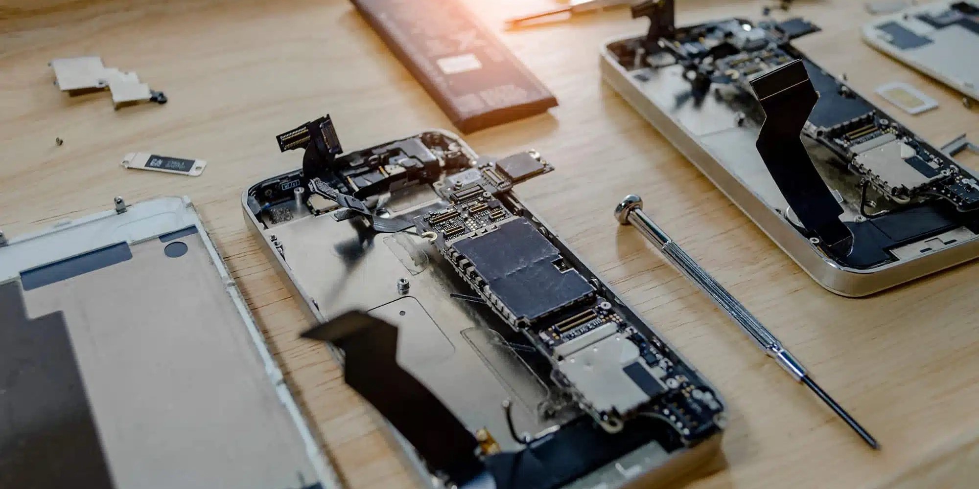 Plusieurs smartphones sont en cours de démontage sur une surface en bois, avec leurs composants internes visibles, notamment des circuits imprimés et des batteries. Un petit tournevis est posé à côté.