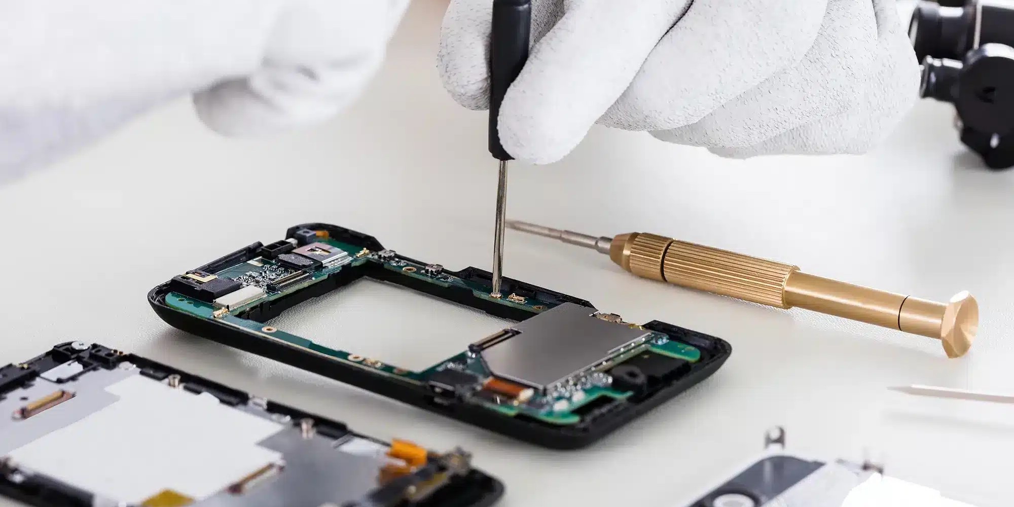Des mains gantées de blanc utilisent un petit tournevis pour réparer un smartphone démonté. La carte mère et d'autres composants sont visibles, avec des outils spécialisés à côté.
