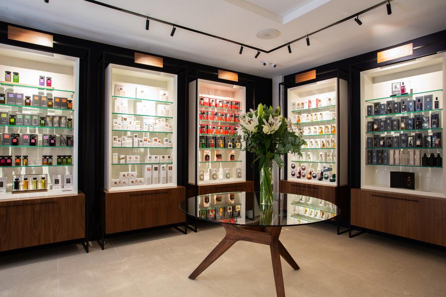 L'intérieur épuré et moderne d'une parfumerie, avec un grand vase de lys blancs sur une table basse en verre au centre. Des vitrines éclairées, remplies de flacons de parfum colorés, ornent les murs, et le plafond est équipé de spots noirs.