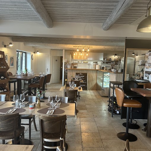 L'intérieur chaleureux et rustique d'un restaurant avec des poutres en bois au plafond. Plusieurs tables en bois sont dressées avec des verres à pied et des serviettes, tandis qu'un bar et une cuisine sont visibles en arrière-plan.