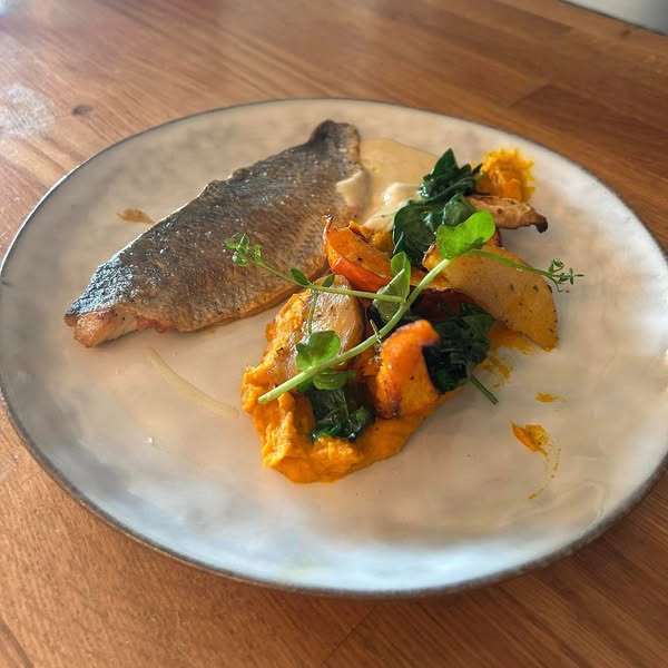 Un plat appétissant est présenté sur une assiette blanche avec un filet de poisson à la peau croustillante. Il est accompagné d'une purée orange, de légumes rôtis comme des carottes et des épinards, et garni de jeunes pousses vertes.