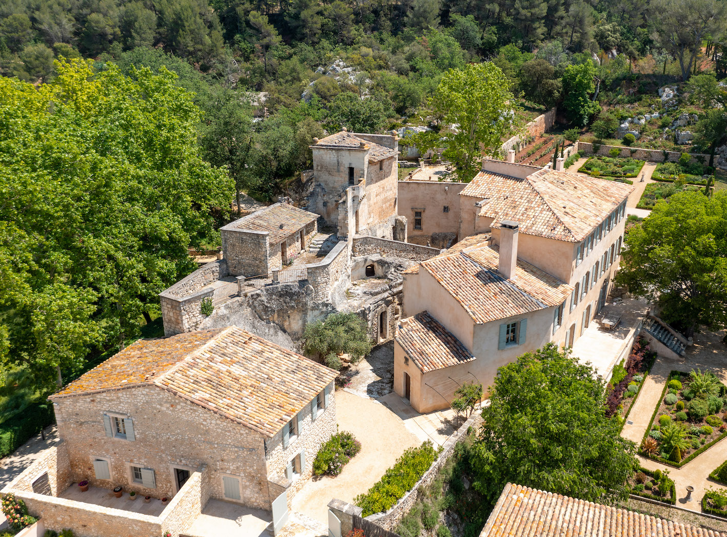 Vue aérienne d'un complexe de bâtiments en pierre aux toits de tuiles, niché au cœur d'une végétation luxuriante et entouré de jardins aménagés.