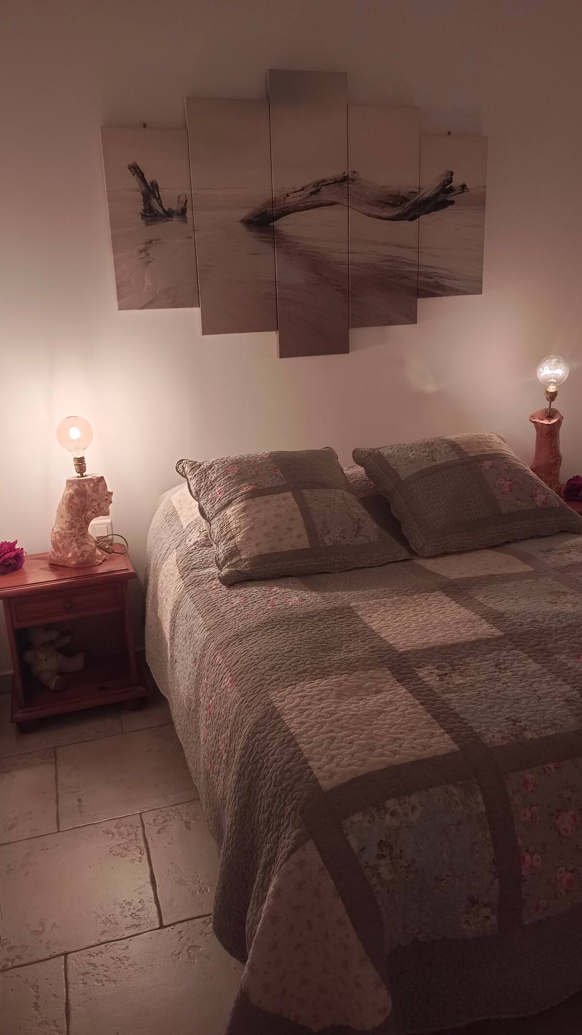 Une chambre douillette avec un lit double orné d'un patchwork, éclairée par deux lampes de chevet au design unique. Au-dessus du lit, un tableau en quatre panneaux représente un morceau de bois flotté sur une plage.