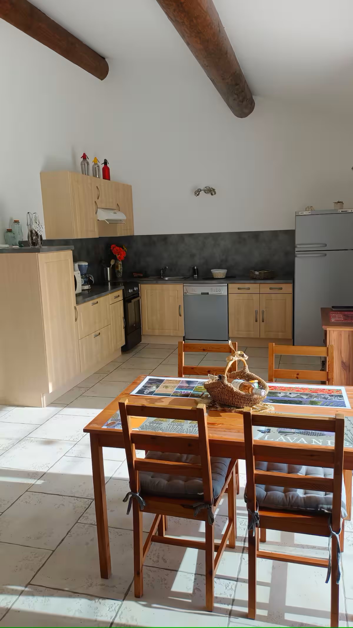 Une cuisine spacieuse et lumineuse avec des meubles en bois clair, un plan de travail sombre et un grand réfrigérateur. Des poutres en bois apparentes traversent le plafond. Au premier plan, une table à manger en bois avec quatre chaises, et un panier de fruits posé sur la table.