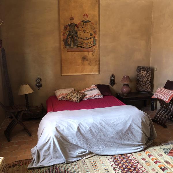 Une chambre rustique avec un grand lit drapé de blanc sur un drap rose, des coussins à motifs, et un tapis persan au sol. Un tableau mural représentant deux figures historiques est accroché au-dessus du lit. Deux lampes de chevet et des objets décoratifs sont visibles, créant une ambiance orientale.