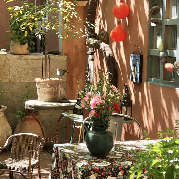 Un charmant patio ensoleillé avec une table recouverte d'une nappe à motifs et un vase de fleurs roses et rouges au centre. Divers objets décoratifs, comme des bouées orange, sont accrochés à un mur de couleur pêche, et des plantes en pot ajoutent une touche de verdure.