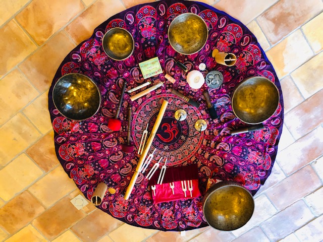 Une composition de bols chantants en cuivre, de diapasons, de maillets et d'autres instruments de méditation est disposée sur un tapis rond à motifs psychédéliques roses et violets, avec le symbole "Om" au centre. Le tout est posé sur un sol carrelé.
