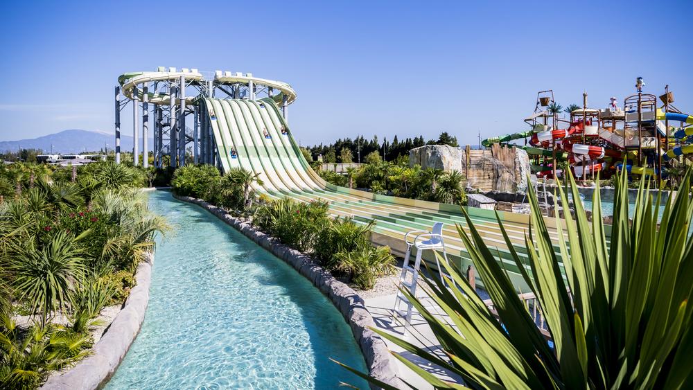 Un grand parc aquatique ensoleillé avec une rivière artificielle aux eaux claires serpentant à travers des plantes tropicales. Plusieurs toboggans massifs, dont un grand toboggan rayé vert et blanc, sont visibles, ainsi qu'une zone de jeux aquatiques colorée en arrière-plan.