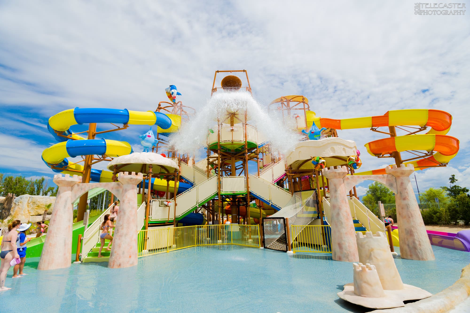 Un parc aquatique animé avec une grande structure de jeux colorée, comprenant des toboggans jaunes, bleus et oranges, ainsi qu'un immense seau déversant de l'eau. Des personnes profitent de l'eau claire sous un ciel partiellement nuageux.