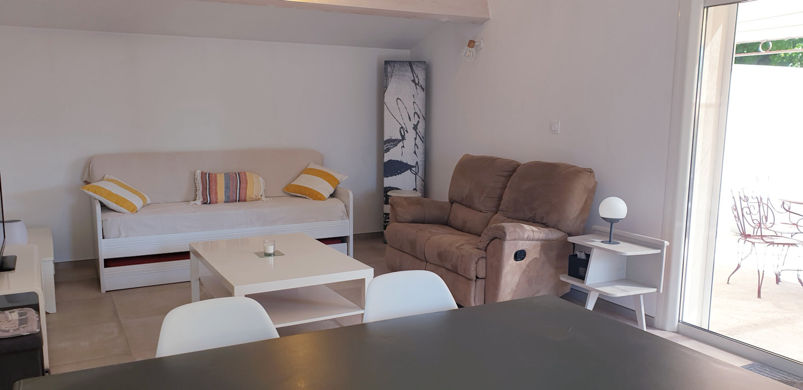 Un salon lumineux avec des murs blancs et un canapé convertible beige agrémenté de coussins colorés. Un fauteuil inclinable marron et une table basse blanche complètent l'ameublement, tandis qu'une grande porte vitrée donne sur l'extérieur.