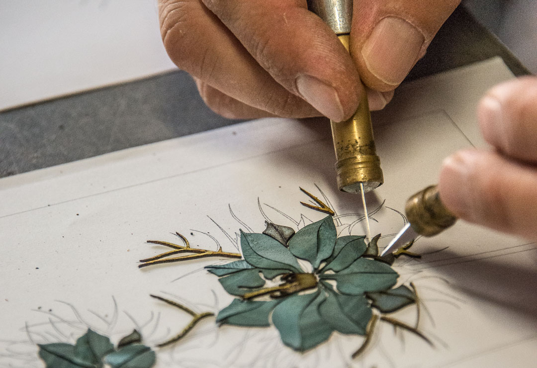Des mains méticuleuses utilisent un outil en laiton pour créer des feuilles en ruban brodé, ajoutant des détails à un motif floral esquissé sur une surface blanche.