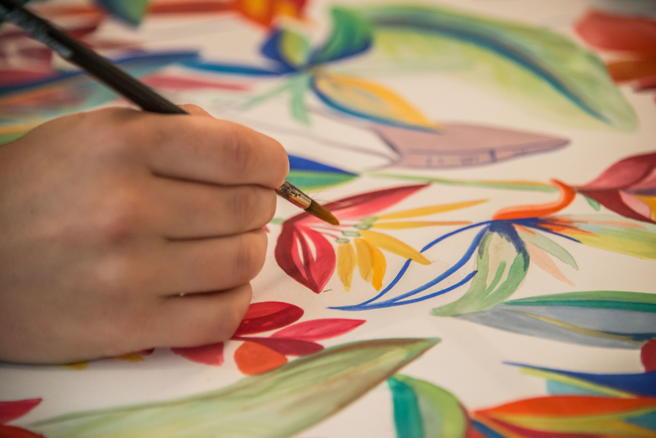 Une main tenant un pinceau est en train de peindre des motifs floraux et de feuilles aux couleurs vives (rouges, jaunes, bleues et vertes) sur une surface blanche, avec des détails précis.