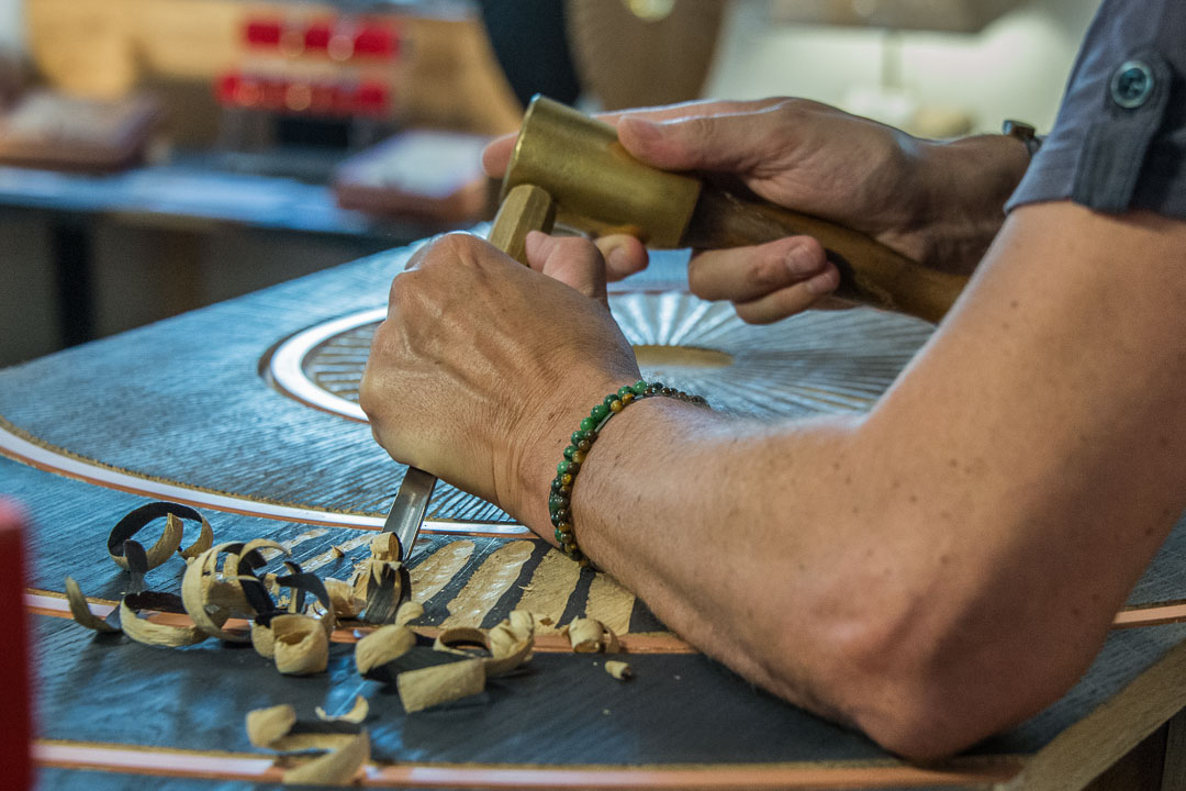 Un artisan travaille sur un projet de marqueterie, utilisant un ciseau et un maillet pour sculpter un motif circulaire sur une surface foncée. Des copeaux de bois clairs s'accumulent autour de ses mains.