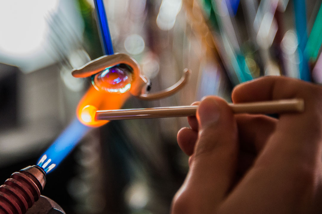 Un souffleur de verre concentré manie une torche à flamme orange et bleue pour sculpter habilement une petite pièce de verre. Ses mains tiennent un outil fin, et des tiges de verre colorées floues sont visibles en arrière-plan.