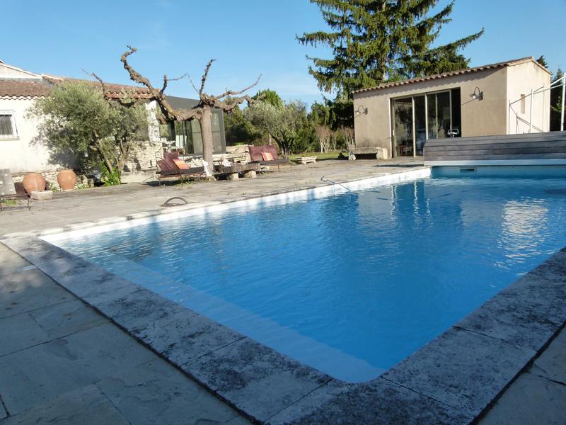 Une grande piscine aux eaux bleues claires est entourée d'une terrasse en pierre et d'un pool house moderne. En arrière-plan, des oliviers et une maison de style provençal contribuent à une ambiance ensoleillée.