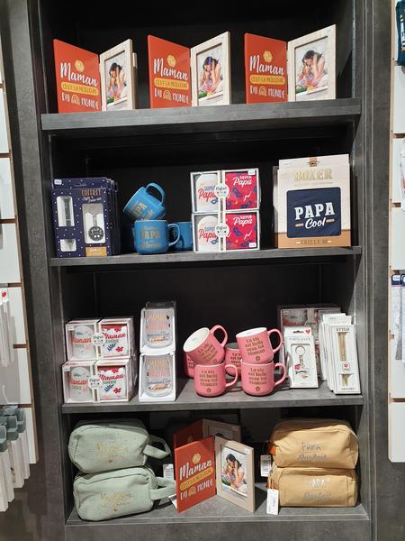 Une étagère de magasin présente des articles cadeaux aux tons de rose, bleu et beige, incluant des tasses, des cadres photo et des pochettes. Plusieurs articles sont marqués "Maman" ou "Papa", et les étagères sont faites de matériaux sombres.