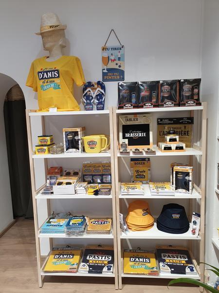 Deux étagères en bois clair sont remplies de produits dérivés à dominante jaune et bleu, comprenant des t-shirts, des mugs, des casquettes et divers articles à l'effigie de marques françaises populaires, notamment le Pastis. Un mannequin porte un chapeau de paille.