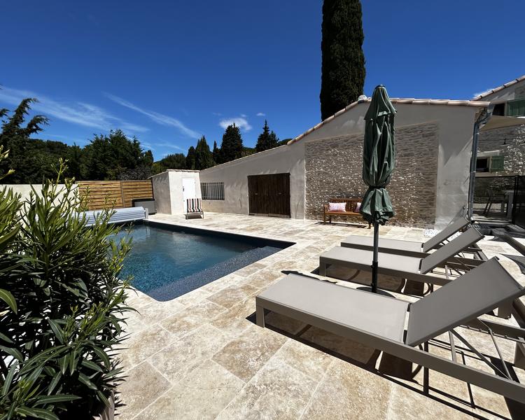 Une élégante terrasse de piscine en pierre, avec une piscine rectangulaire aux eaux bleues, des chaises longues et un parasol, le tout sous un ciel bleu clair.