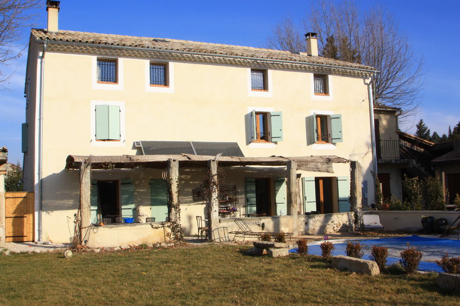 Une grande maison provençale à trois étages avec des volets bleus et un auvent, un jardin, une piscine recouverte et un ciel bleu.