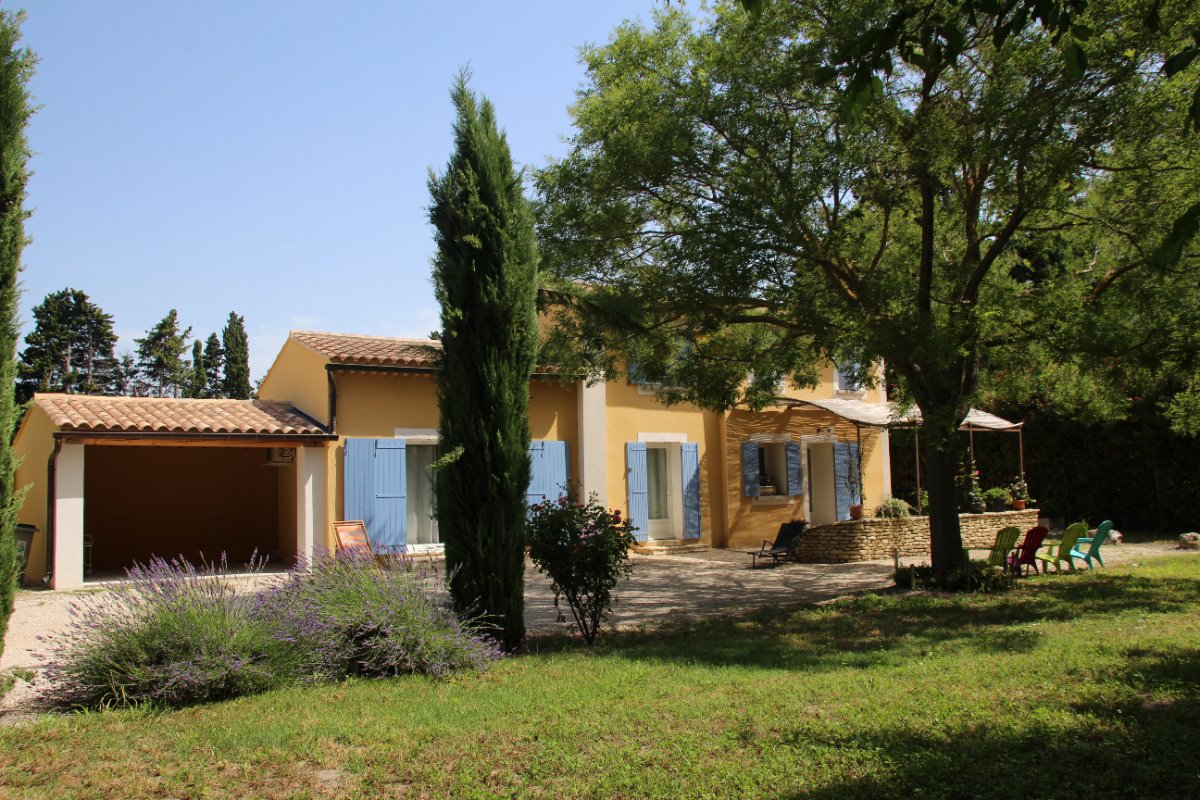 Une maison provençale jaune avec des volets bleus, un garage, et un grand jardin verdoyant avec des arbres, de la lavande et des chaises colorées.