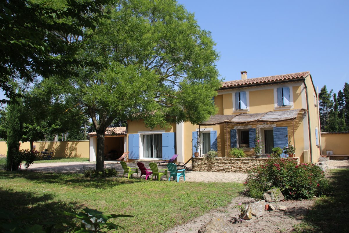 Une charmante maison provençale jaune avec des volets bleus, entourée d'un grand jardin verdoyant avec des arbres, de la lavande et des chaises de jardin colorées.