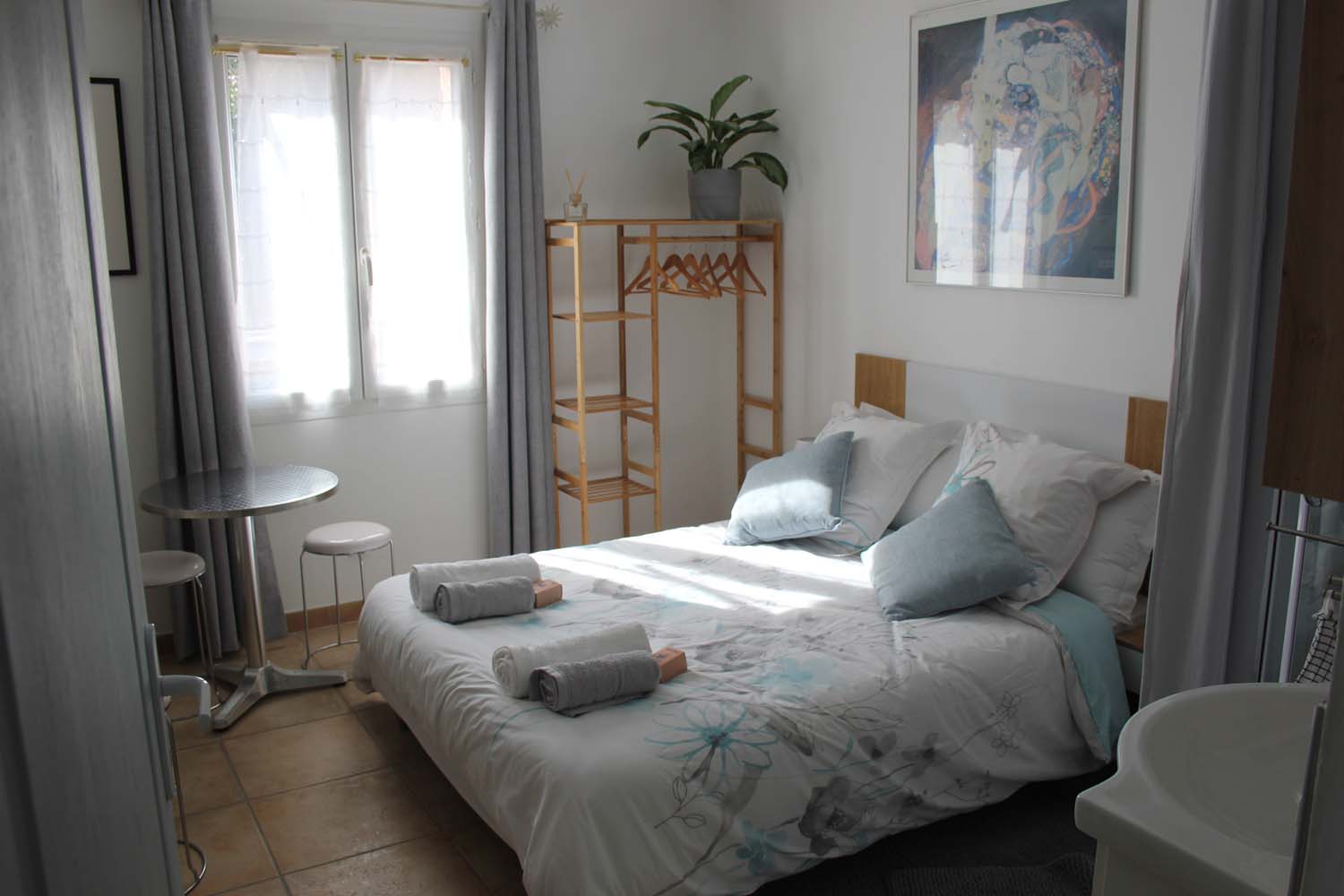 Une chambre lumineuse et moderne avec un lit double, une table ronde avec des tabourets, une penderie en bois et une plante verte, sous une lumière naturelle éclatante.