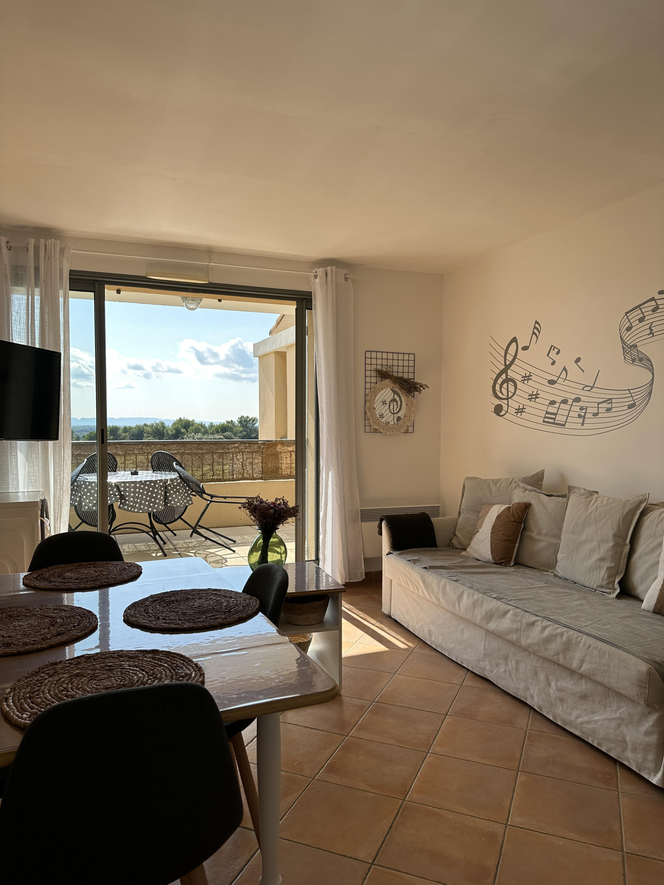 Un salon lumineux avec une table à manger, un canapé, une décoration murale musicale, et une porte-fenêtre donnant sur une terrasse ensoleillée avec vue.