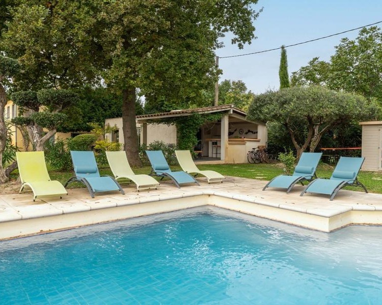 Une piscine extérieure avec des chaises longues colorées, un pool house rustique et de la verdure en arrière-plan sous un ciel clair.