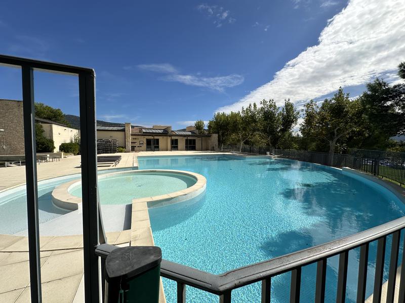 Une grande piscine extérieure avec un bain à remous intégré, entourée d'une terrasse et de verdure, sous un ciel bleu parsemé de nuages.