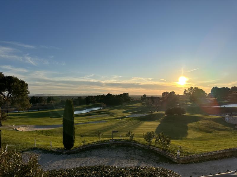 Un parcours de golf verdoyant avec des points d'eau, des arbres et des bunkers, sous un coucher de soleil éclatant.