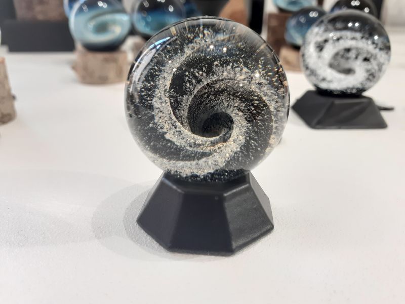 Une sphère en verre sur un socle noir, contenant un motif tourbillonnant blanc et gris ressemblant à une galaxie, avec d'autres orbes en arrière-plan.