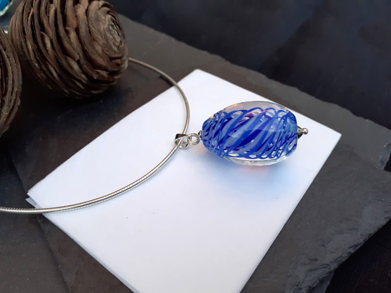 Un collier avec un pendentif en verre bleu et blanc, reposant sur une surface sombre avec des pommes de pin en arrière-plan.
