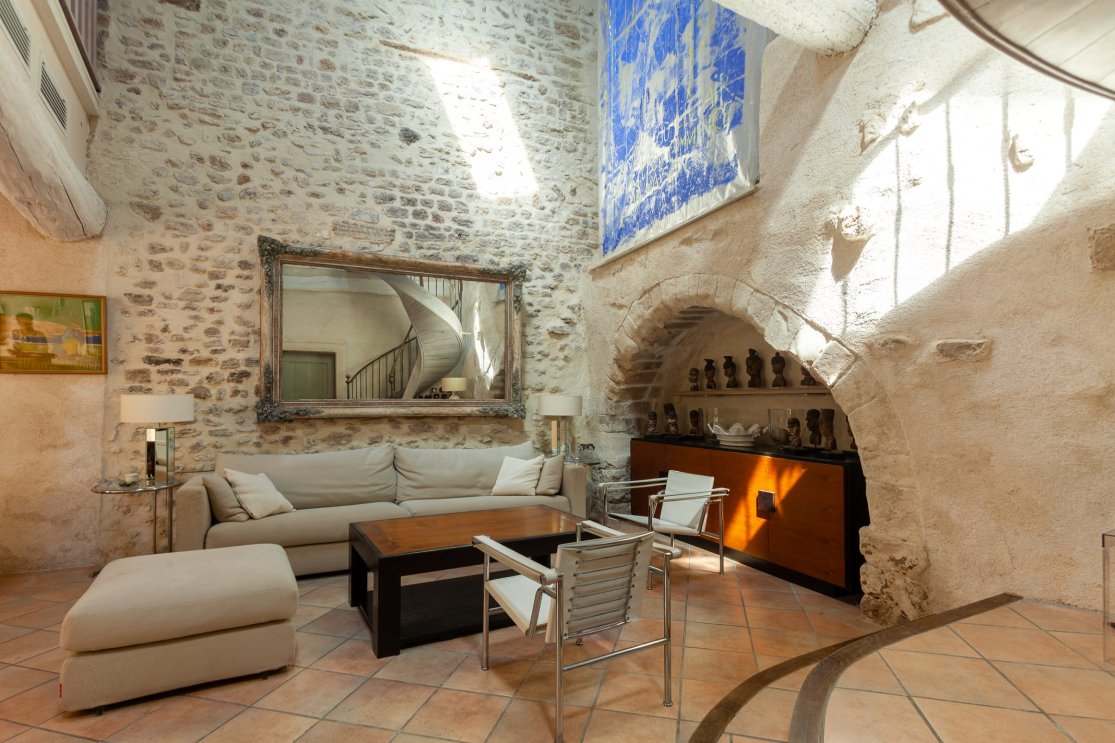 Un salon spacieux avec des murs en pierres apparentes, une grande œuvre d'art abstraite bleue accrochée en hauteur, un canapé beige avec des coussins, deux fauteuils modernes avec des cadres métalliques, une table basse en bois, un pouf rembourré, et un miroir encadré de façon complexe reflétant un escalier en colimaçon. Une niche encastrée dans le mur est remplie d'objets décoratifs, et le sol est carrelé de terre cuite. La pièce est bien éclairée par la lumière naturelle.