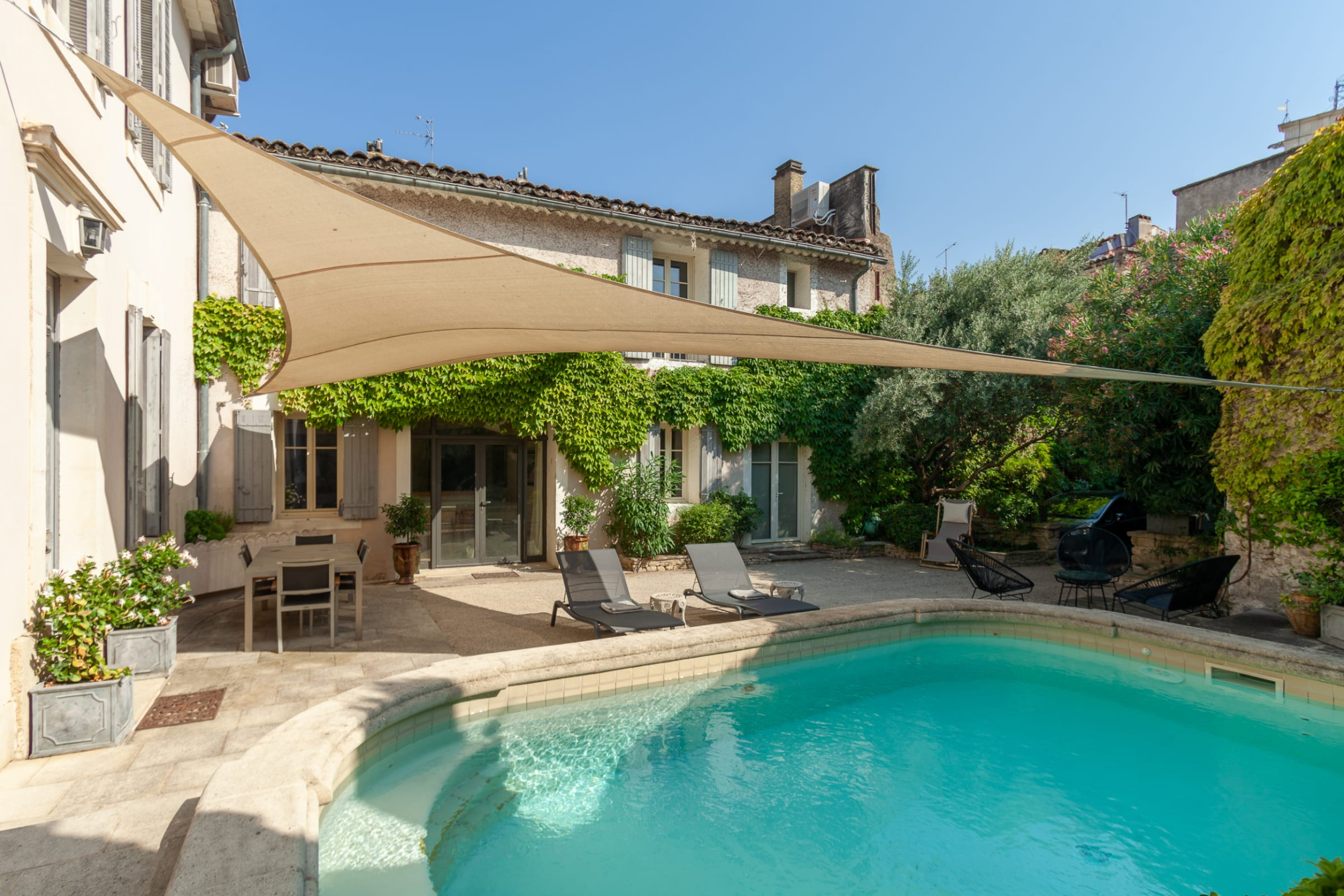 Une piscine extérieure en forme de haricot avec de l'eau bleue claire, entourée d'une terrasse en pierre et d'un jardin luxuriant. Des chaises longues sont disposées au bord de la piscine sous un voile d'ombrage beige. Un bâtiment traditionnel avec des volets et de la vigne grimpante est visible en arrière-plan.
