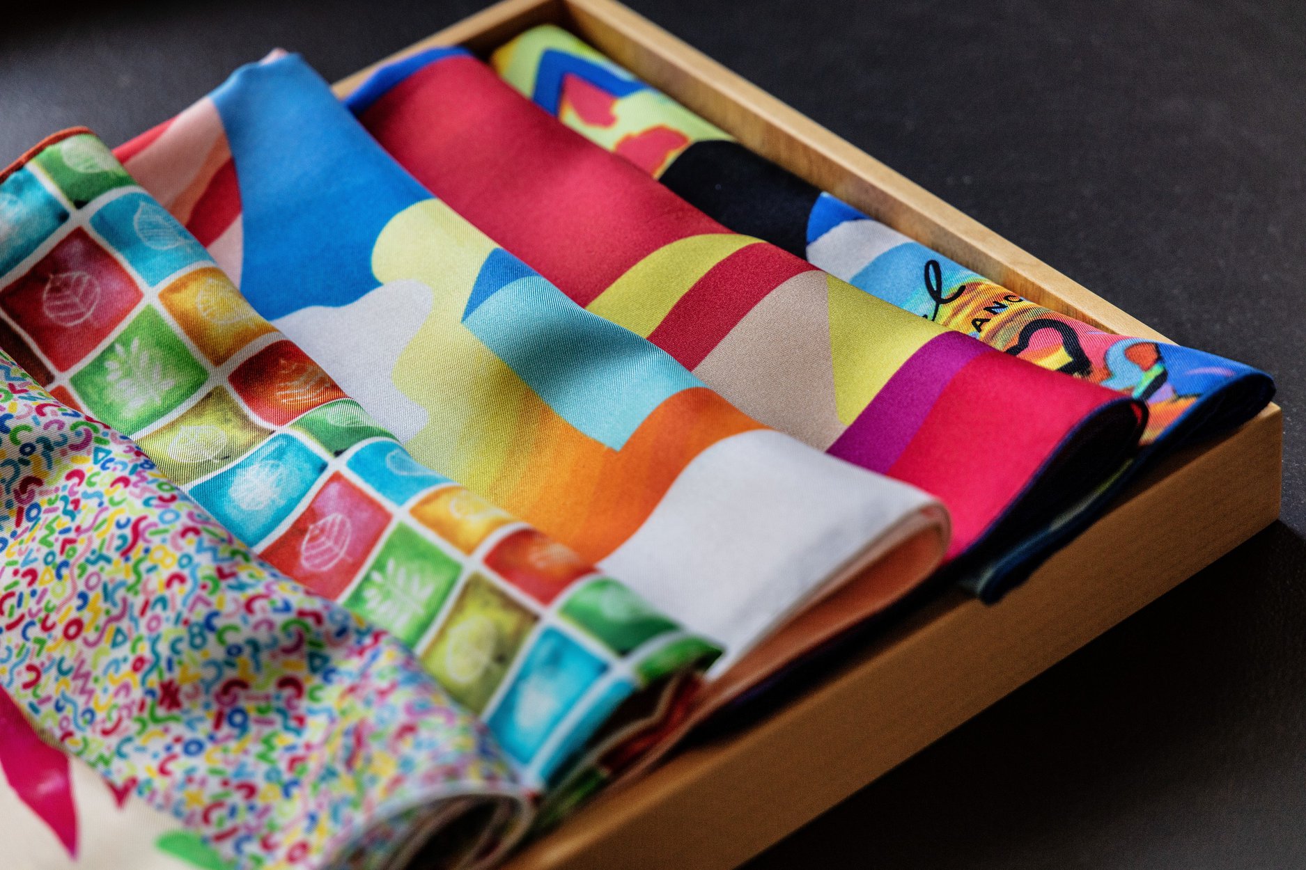 Une boîte en bois remplie de mouchoirs ou de foulards en tissu aux motifs et couleurs variés et vibrants, certains avec des dessins géométriques et d'autres abstraits.