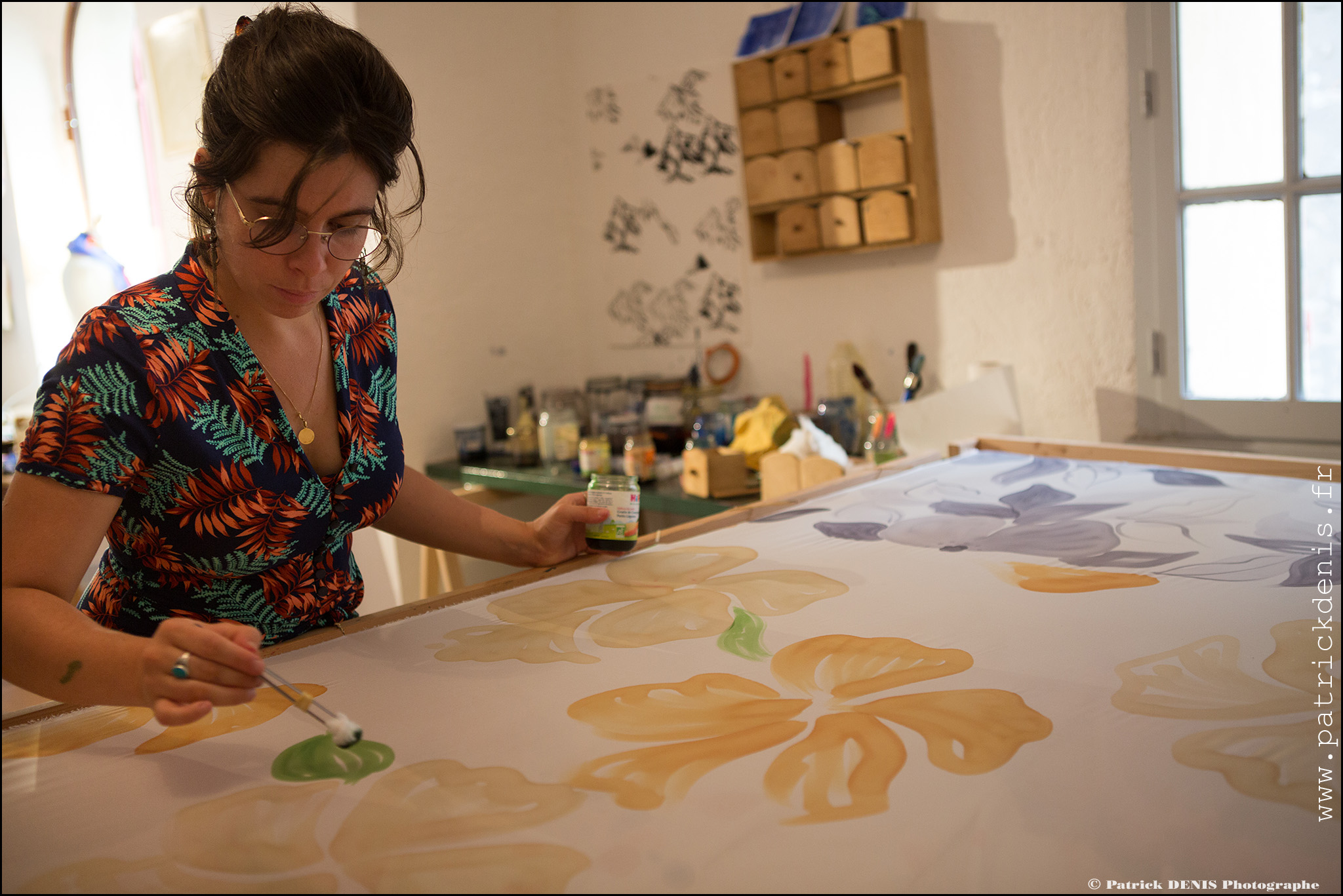 Une artiste concentrée, peignant des motifs floraux sur une grande pièce de tissu blanc, dans un atelier lumineux.