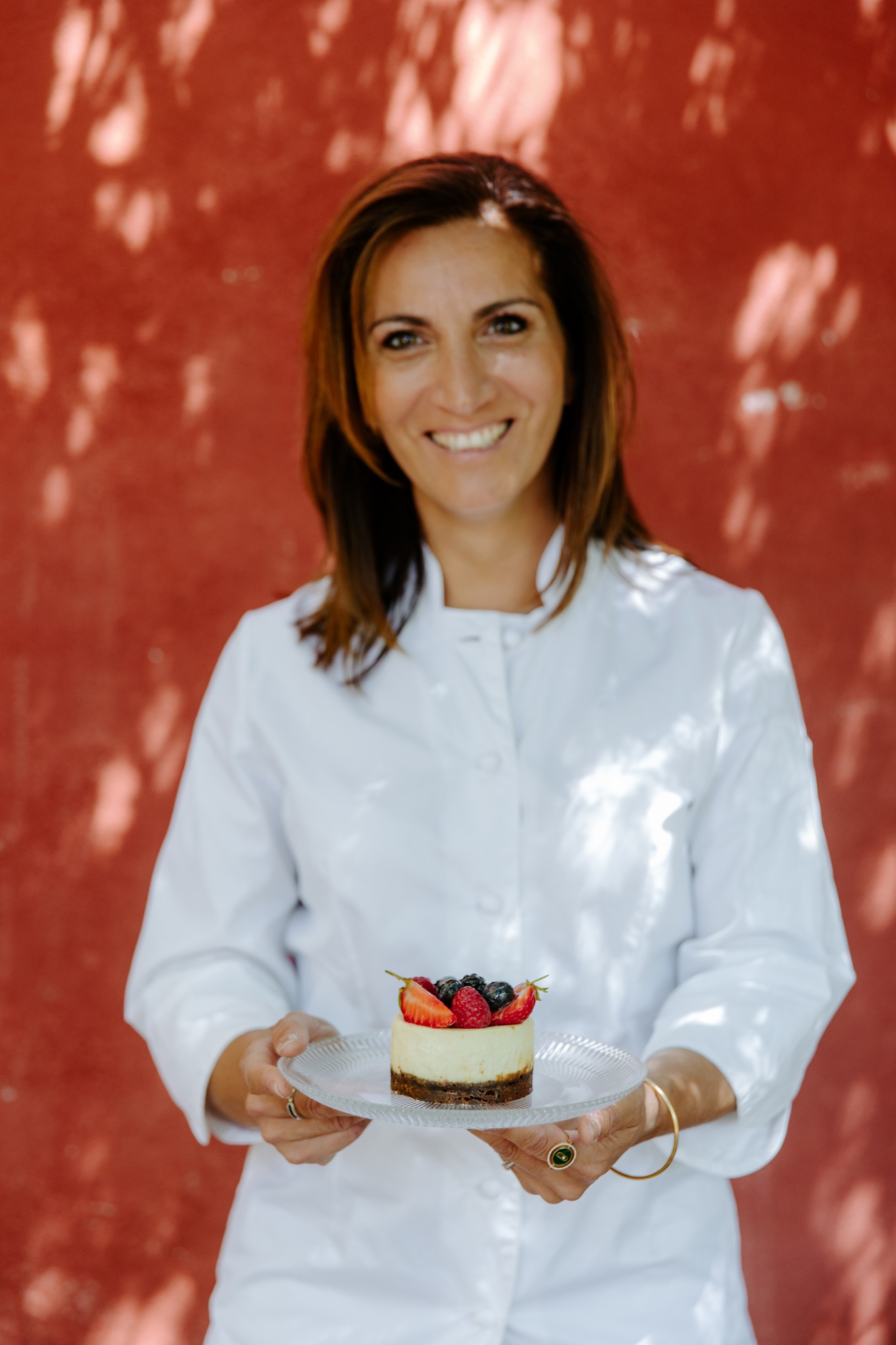 Une cheffe pâtissière souriante vêtue d'une toque blanche et d'une veste de cuisine, tenant un cheesecake individuel garni de baies rouges et bleues sur une assiette en verre.