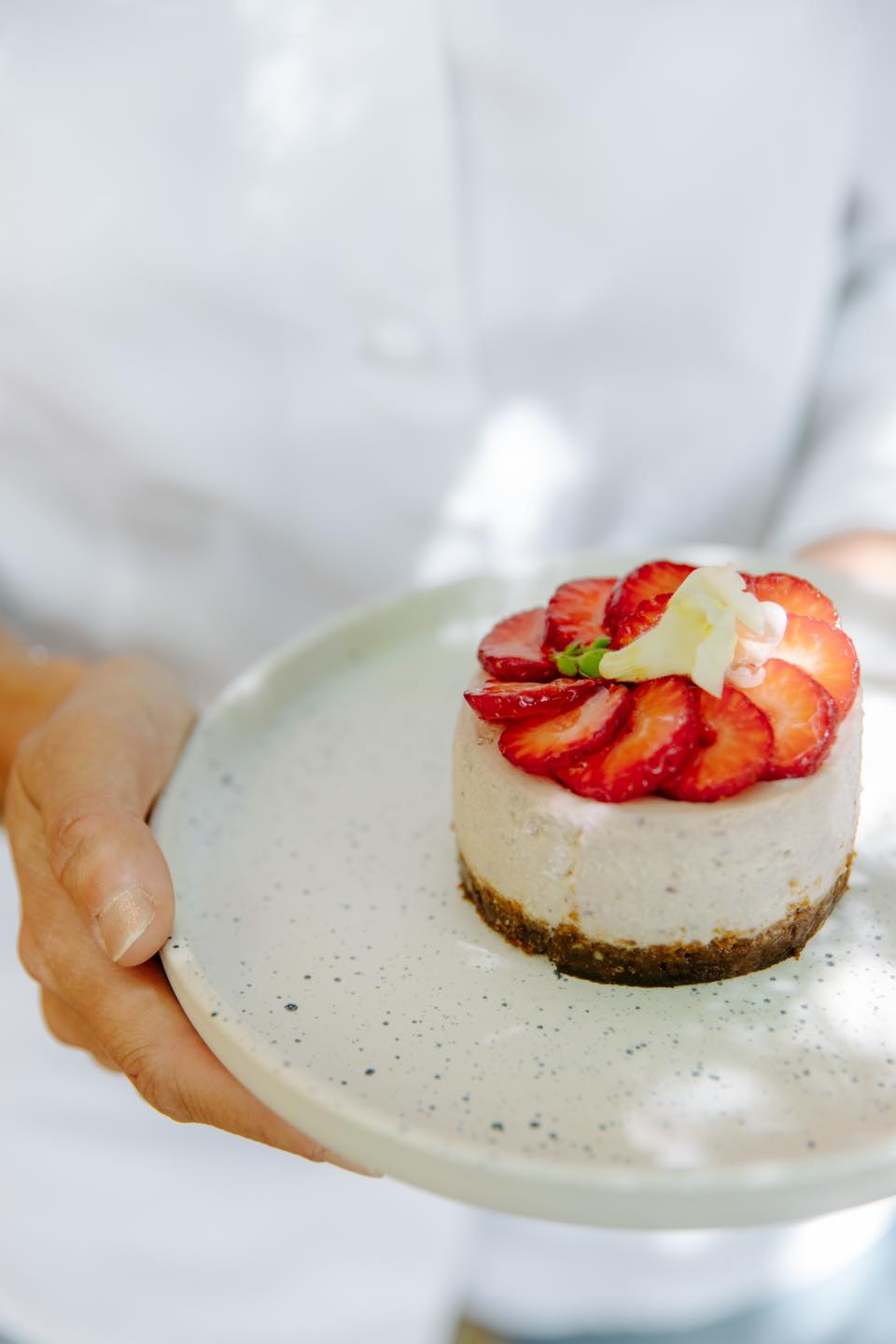 Une personne portant une chemise blanche tient une assiette blanche avec un mini cheesecake garni de fraises tranchées et d'une petite fleur blanche.